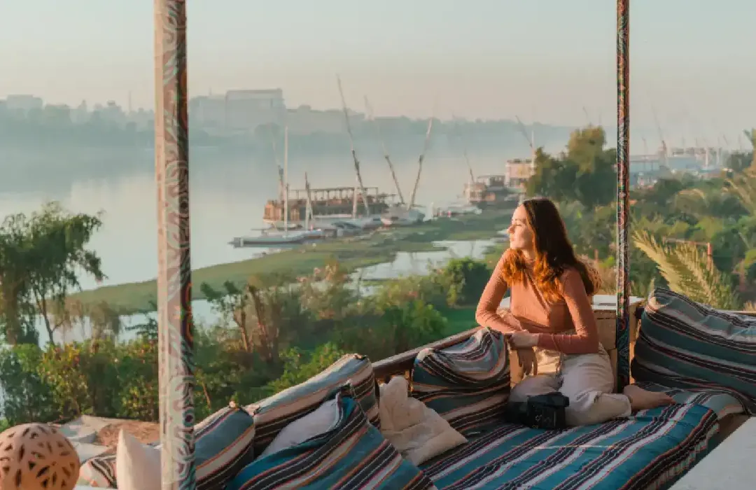 Une femme assise sur une terrasse avec des coussins rayés, admirant le Nil